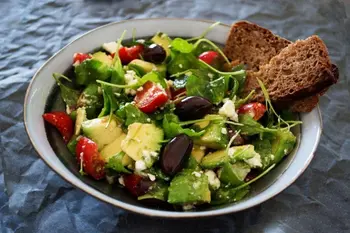kase içinde zeytin, salatalık,roka,domates ve peynir bulunan bir salata. Kenarında kızartılmış ekmekle servis edilmiş. ÜZerinde zeytinyağı gezdirilmiş. Lezzetli görünüyor.